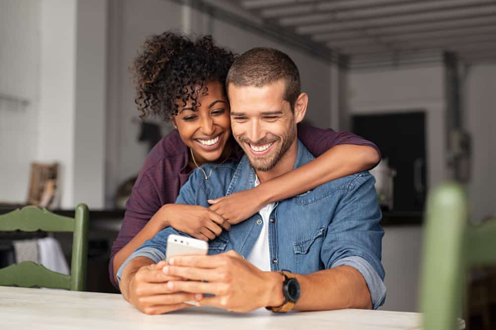 A female and male embracing while looking at a smartphone.